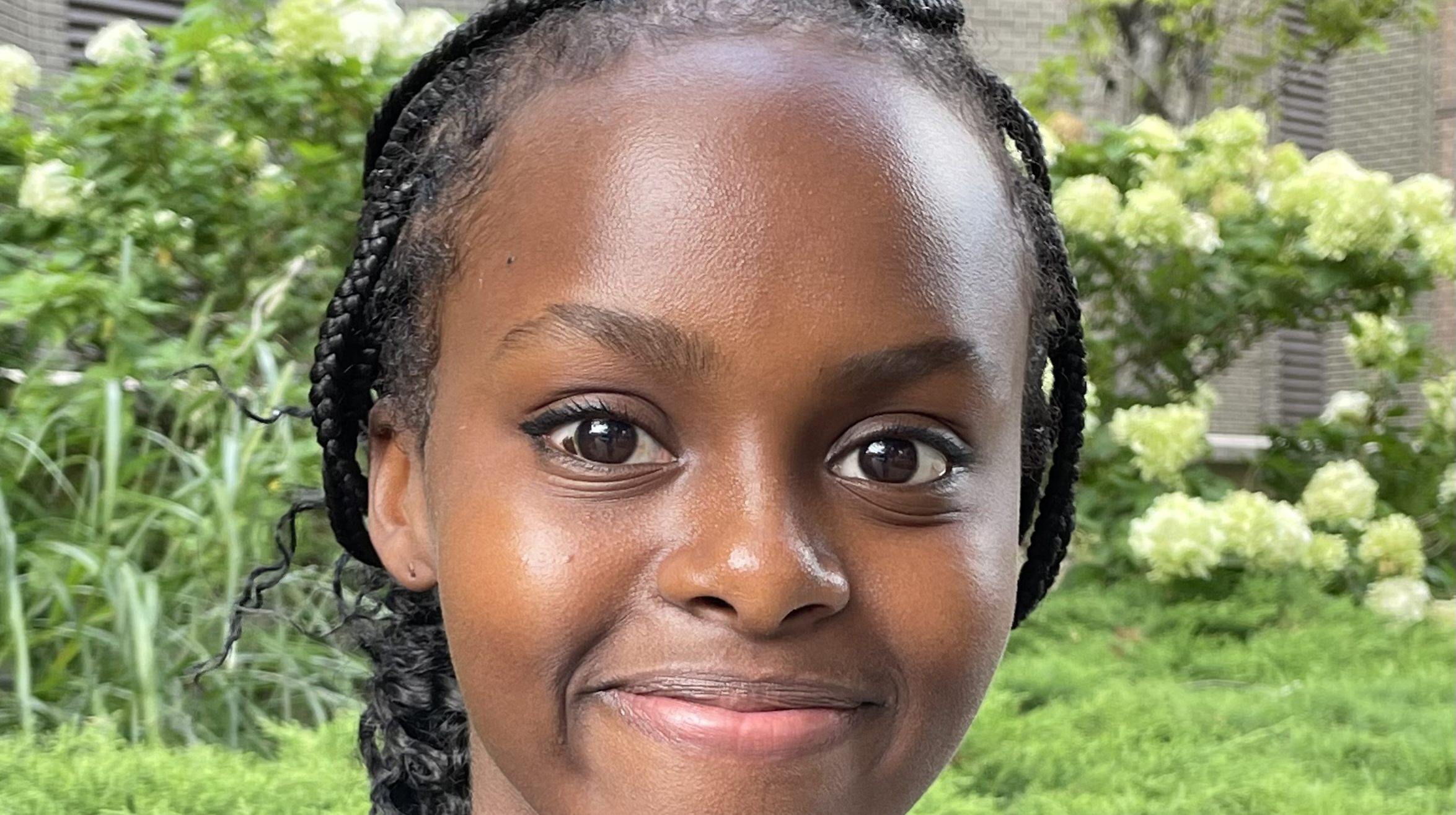 Portrait of a current Gannon Scholar with braided hair pulled back, smiling softly, photographed outdoors with green plants and light-colored flowers in the background.